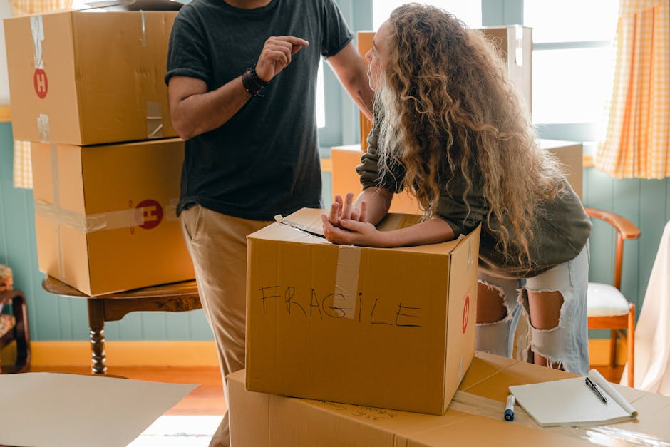 A person wearing a black t-shirt and beige trousers is standing inside a room, engaging in a home relocation process with a woman who has curly blonde hair, dressed in a grey long-sleeve top and ripped white jeans. The woman is leaning over a cardboard box labeled 'FRAGILE' with handwritten black marker, and appears to be inspecting or preparing to pack it. The scene includes several stacked cardboard boxes, some sealed with packing tape and bearing printed red symbols, situated on a wooden table and on the floor. The room features green paneled walls, a window with orange checkered curtains allowing natural light to fill the space, and some furniture such as a white chair with orange armrests visible in the background. The environment suggests a packing and loading process typical of furniture transport or home relocation, with attention to careful handling of fragile items as part of the moving services of Man With a Van Eltham.