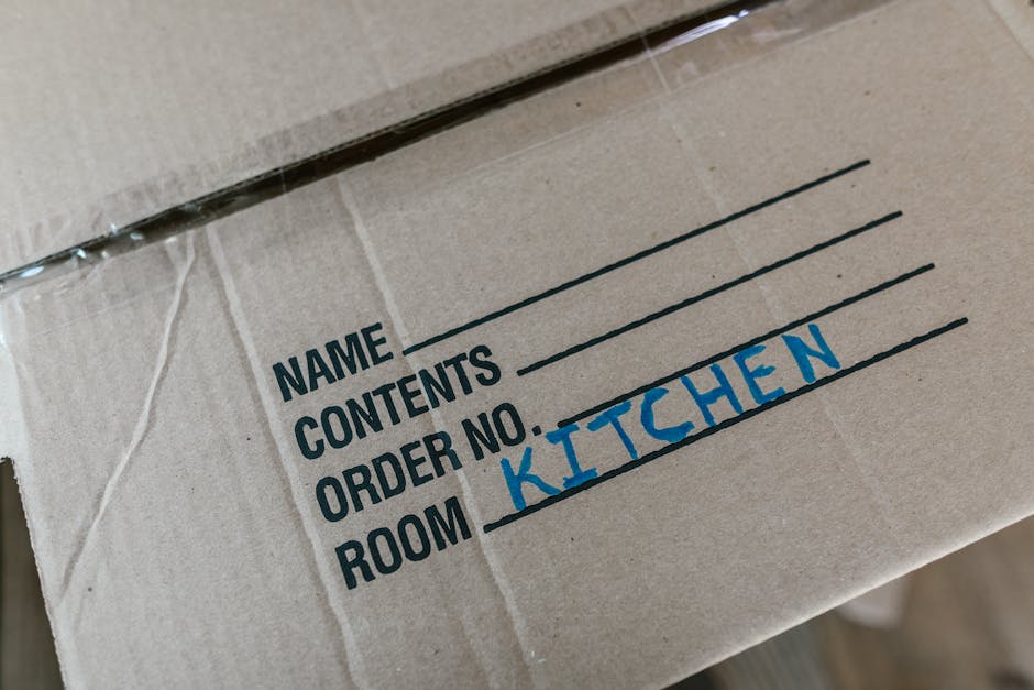 Close-up of a cardboard moving box with a focus on the printed label sections. The box has handwritten and printed details, revealing a section for the recipient's name, contents, and order number. The handwritten part indicates the contents as 'KITCHEN' in blue ink, and the box appears to be sealed with clear packing tape. The background shows a glimpse of other boxes or packing materials, suggesting a home relocation or packing process. This type of packaging is typical during house removals facilitated by Man With a Van Eltham, emphasizing organized packing and furniture transport as part of the moving services.