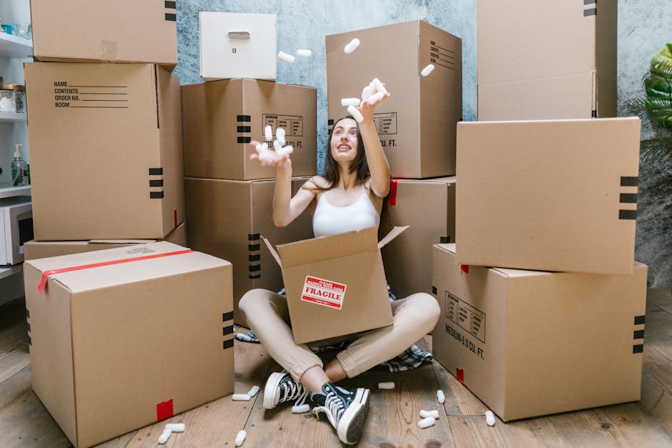 A young woman sitting cross-legged on a wooden floor inside a room surrounded by packed cardboard boxes of various sizes, some with labels and red tape, during a home relocation process. She is smiling and tossing white packing foam into the air while holding a box marked with a 'Fragile' sticker, indicating careful handling. The boxes are stacked against a textured wall, and the room contains a small potted plant and a piece of furniture in the background. The scene captures the packing and moving logistics involved in house removals, with a focus on the packing materials and organized boxes ready for transport, as managed by Man With a Van Eltham.