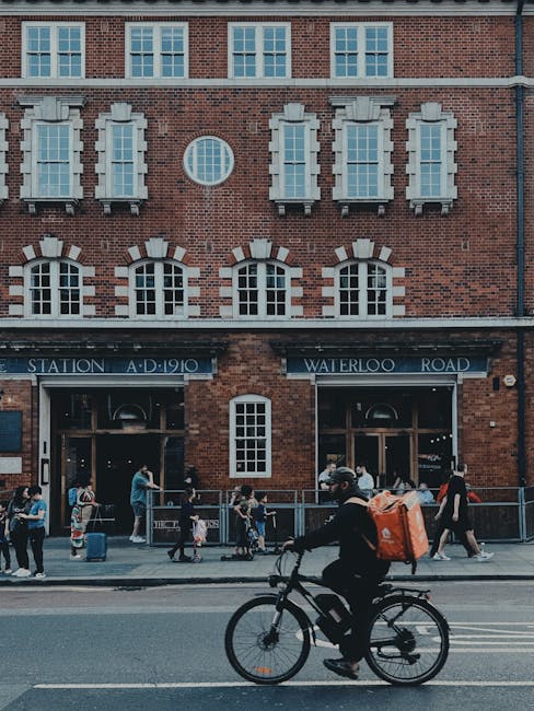 A cyclist riding a black bicycle along a street in front of a historic brick building with multiple large windows and decorative stonework, including a circular window and white window surrounds. The building features a blue sign with white lettering indicating 'Station A.D. 1910' and 'Waterloo Road,' suggesting it is a railway station or station building. Several pedestrians, including adults and children, are walking along the pavement near the building, some carrying bags or pushing strollers. The scene is set during daytime with overcast lighting, and the environment reflects an urban area suitable for home relocation or moving services. The image captures the busy atmosphere typical of transport hubs, supporting services like removals and furniture transport provided by Man With a Van Eltham.