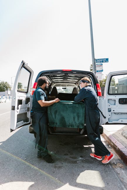Two professional movers from Man With a Van Eltham are engaged in a home relocation process, carefully loading a large, dark green upholstered piece of furniture into the back of a white van parked on a paved street. The interior of the van is visible with partially open side doors, revealing the spacious cargo area designed for transporting bulky items during packing and moving activities. The movers are dressed in dark uniforms, wearing gloves for safety, with one mover guiding the furniture from inside the van while the other supports it from outside. The scene takes place outdoors on a bright day, with a street sign visible on a pole nearby, emphasizing the urban setting of the move, which involves loading furniture into the vehicle for safe transport to a new residence. This image captures an essential stage of the furniture transport and logistics involved in local house removals, demonstrating the careful handling and organization typical of professional moving services provided by Man With a Van Eltham.