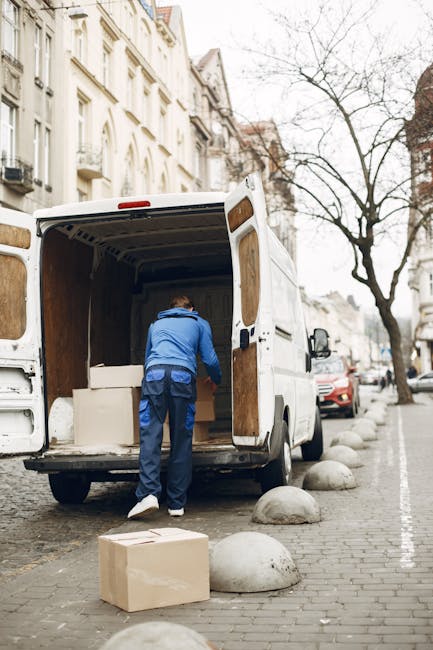 A man dressed in a blue jacket and dark trousers is loading a large cardboard box into the back of a white moving van parked on a cobbled street in an urban residential area. The van's rear doors are 