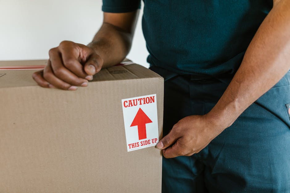 A man dressed in a blue jacket and dark trousers is loading a large cardboard box into the back of a white moving van parked on a cobbled street in an urban residential area. The van's rear doors are 