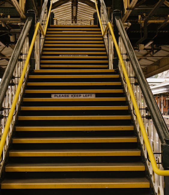 A set of steep, yellow and black striped industrial stairs with metal handrails on both sides, leading up inside a multi-level facility or warehouse. The steps are made of durable material, with some