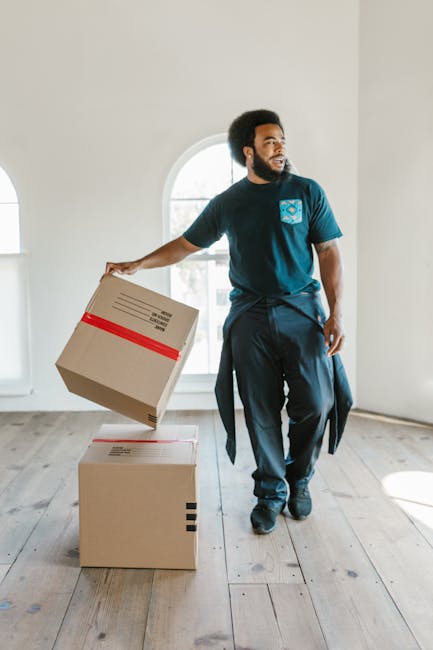 A man with dark curly hair and a beard is inside a bright, empty room with light-colored wooden flooring and large arched windows. He is dressed in a blue t-shirt with a logo on the chest, black trousers, and black shoes. The man is holding a cardboard box with red tape on top in his right hand, preparing to place it on a stack of two similar cardboard boxes on the floor. One box is horizontal on the wooden surface, while the other is upright, ready for stacking during the home relocation or furniture transport process. The room is well-lit with natural daylight streaming through the windows, casting soft shadows on the floor. The environment is minimal, with no furniture or decorations, highlighting the packing and moving preparations often associated with house removals and moving services offered by companies like Man With a Van Eltham.