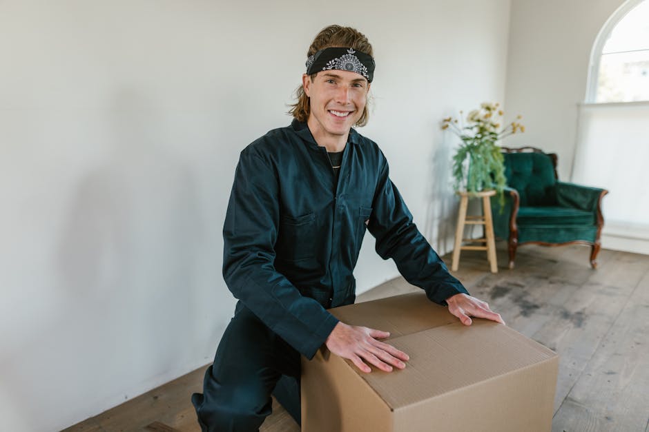 A professional removalist from Man With a Van Eltham is inside a well-lit living room, lifting or preparing to transport a medium-sized cardboard box. The man, dressed in a black uniform with a black headband, is smiling and appears ready for a home relocation process. The room has wooden flooring and features a green upholstered armchair with carved wooden legs positioned near a large window, which allows natural light to illuminate the space. Behind the man, there is a small wooden side table with a potted plant and white flowers, adding to the tidy interior. The scene captures the packing and moving stage, with the removalist handling packing materials carefully and demonstrating the logistics involved in furniture transport and home removals. This image exemplifies professional packing and moving practices as part of a well-planned house removal, consistent with the services offered by Man With a Van Eltham for narrow access moves, emphasizing safe handling of belongings and efficient loading procedures.